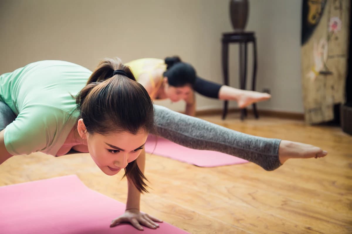 Vrouw zit in kleermakerszit op een mat, terwijl op de achtergrond iemand een yogahouding aanneemt, ter illustratie van het verschil tussen yoga, meditatie en mindfulness